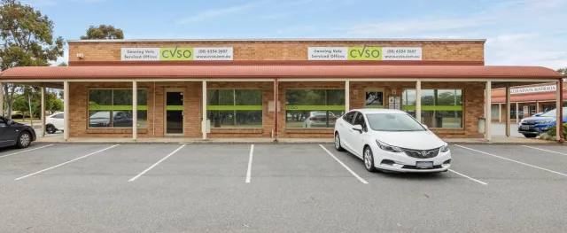 Bright interior photo showing a furnished serviced office in a suburban coworking building: warm timber desks, neutral upholstery, plants, and natural daylight from large windows; photorealistic and inviting.