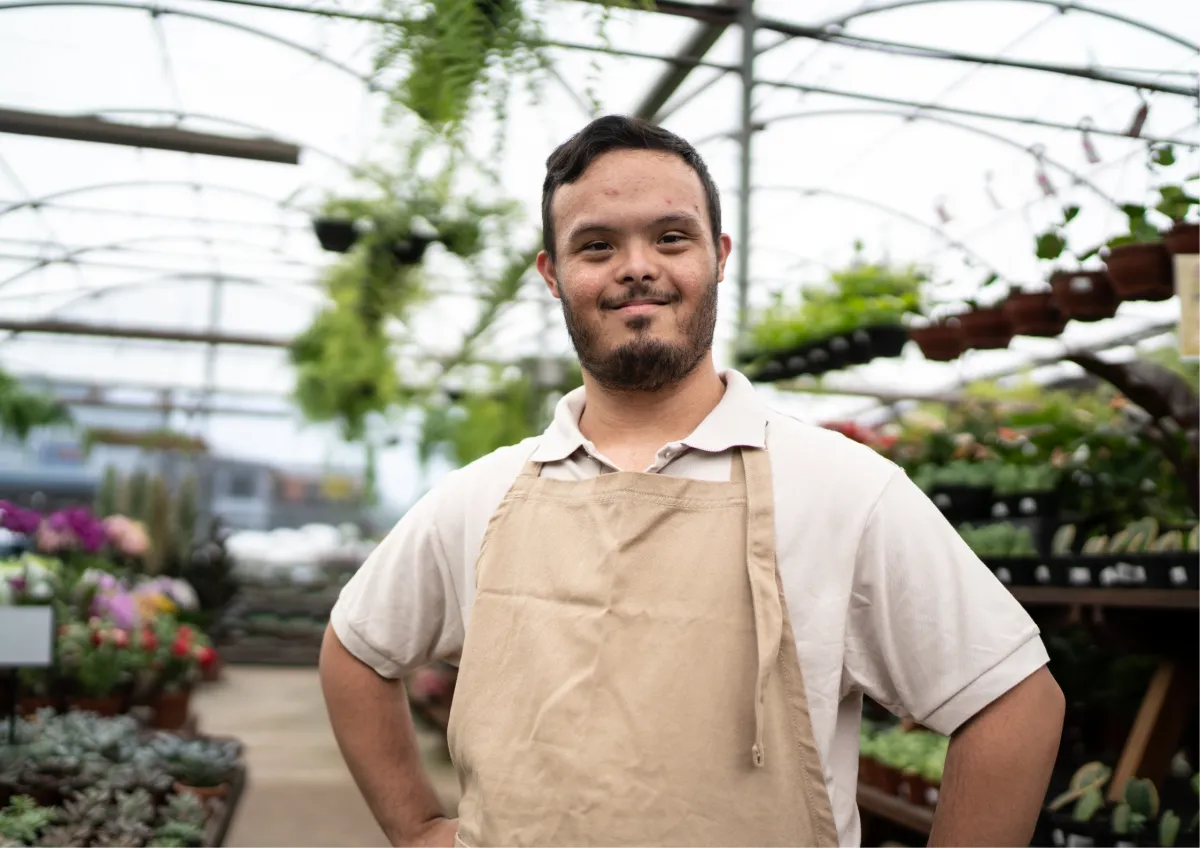 A smiling young man with Down syndrome wearing a tan apron and standing confidently in a garden center greenhouse, highlighting successful inclusive hiring and supported employment in the community.
