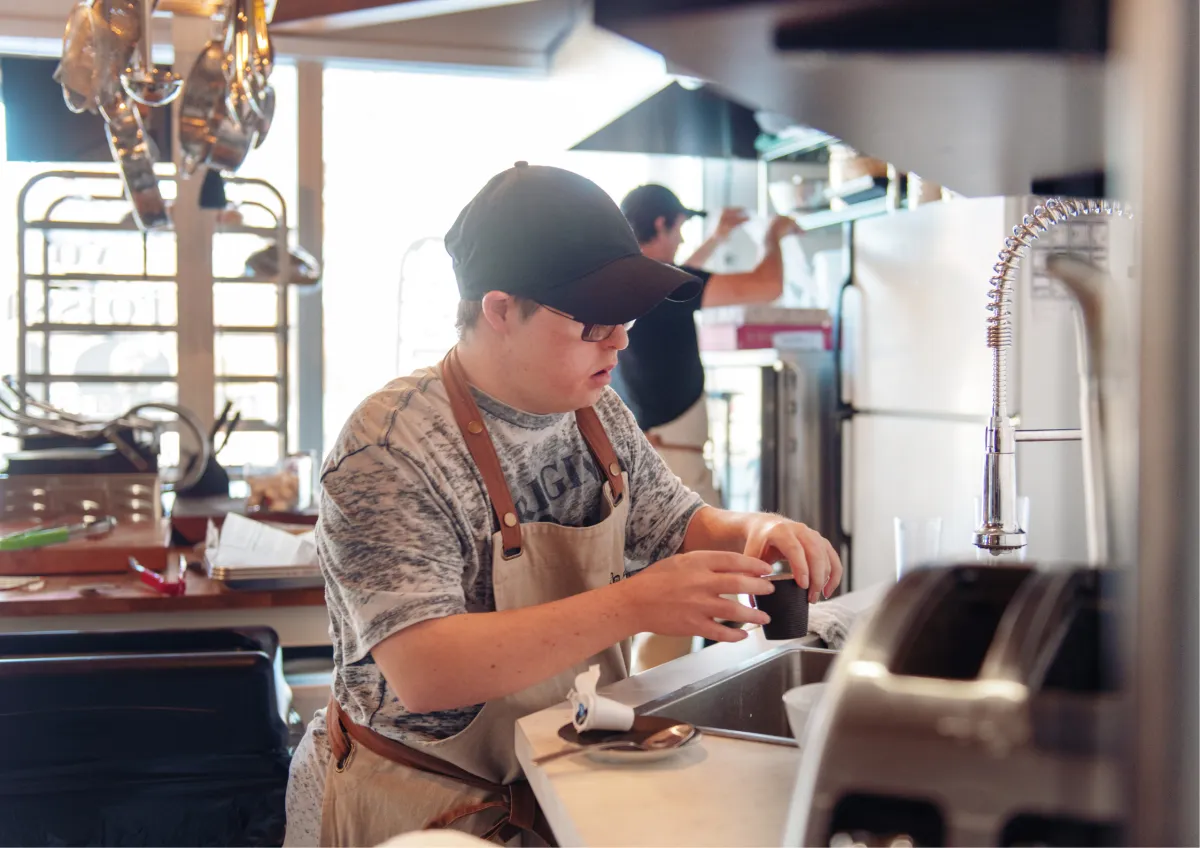 A young male employee with Down syndrome wearing an apron and cap, carefully preparing a drink in a professional kitchen or cafe setting.