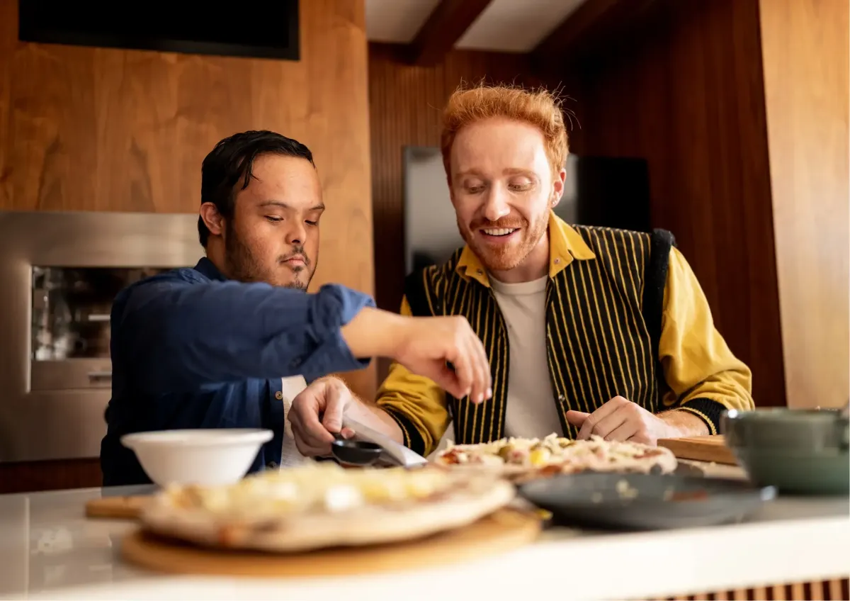 Two friends laughing and having fun while preparing homemade pizza in a modern kitchen.