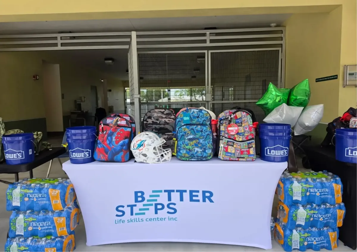 Better Steps Foundation display table with Spider-Man and Super Mario backpacks, Lowe's buckets, and a signed Miami Dolphins helmet