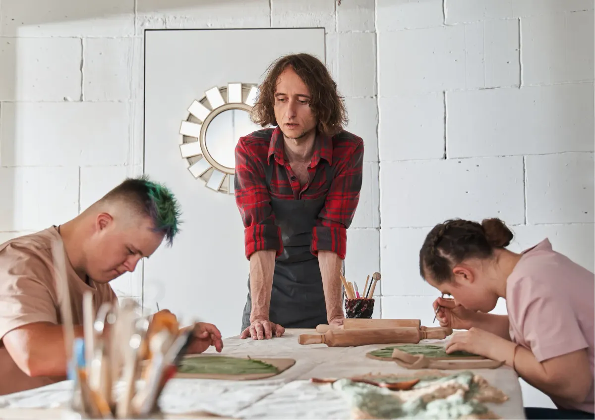 Instructor guiding two adults as they shape clay projects during a pottery class
