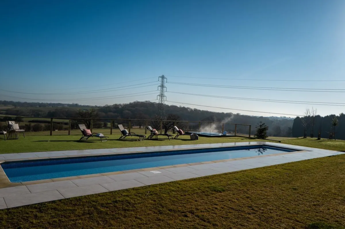 Rectangular pool with stone tiles, loungers on grass, and a steaming raised hot tub overlooking rolling countryside.