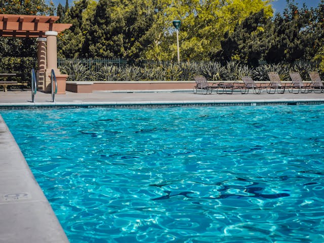 Outdoor swimming pool with clear blue water, poolside loungers, and a pergola surrounded by trees and greenery.