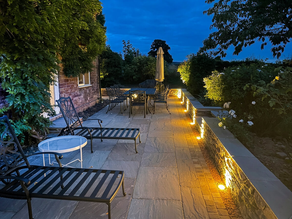 Evening garden patio with stone paving, warm wall lighting, black metal furniture, and surrounding plants beside a brick house.