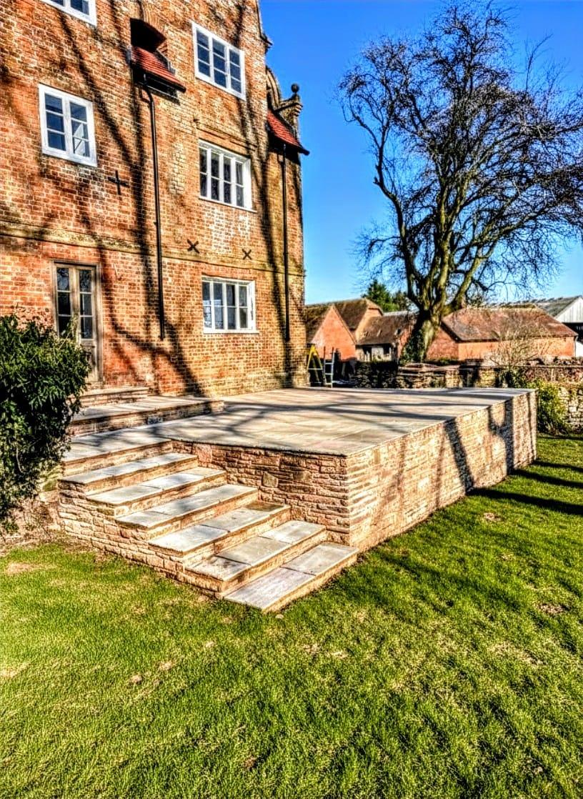 Raised stone patio with wide steps leading to a lawn, set against a traditional brick house in bright sunlight.