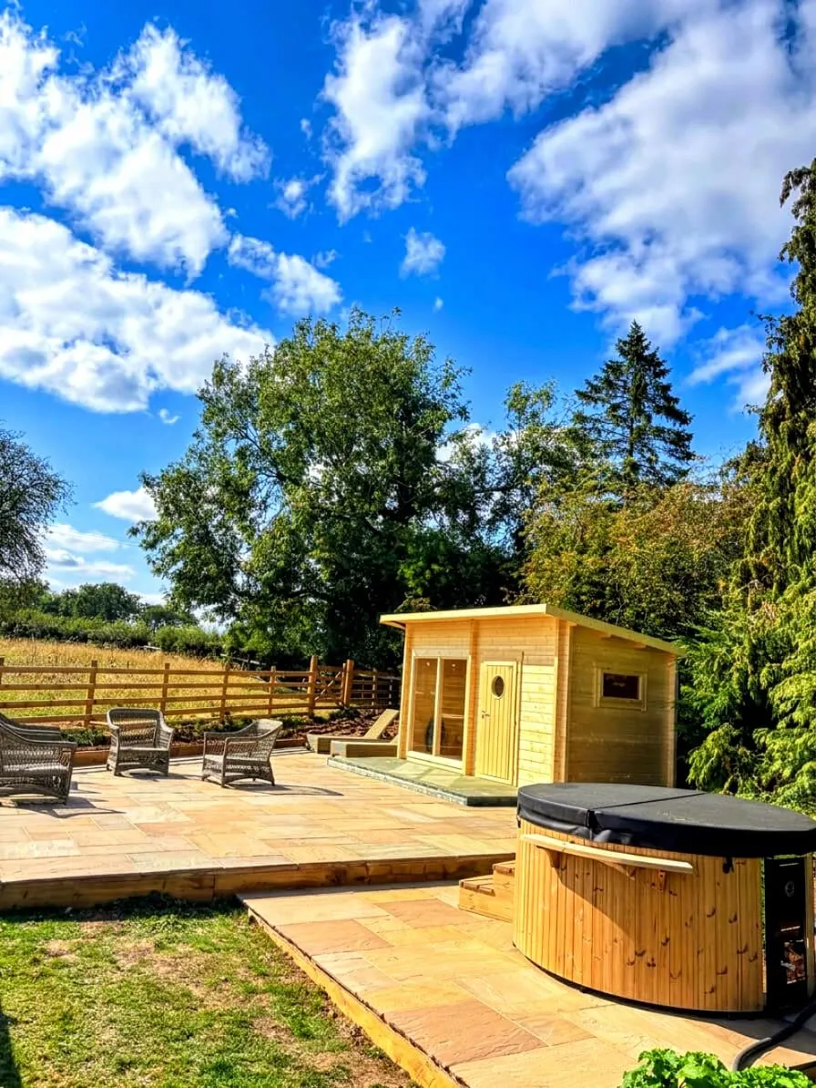 Garden patio with wicker seating, wooden hot tub, and summer garden house set against fields and trees under a bright sky.