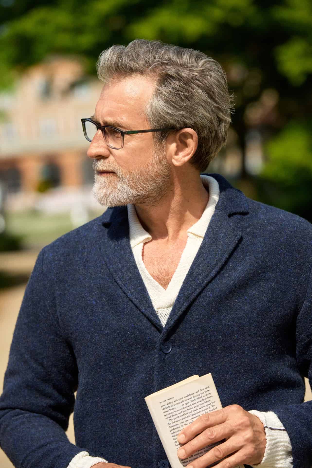 Mature man wearing modern eyeglasses and a navy jacket while holding a book outdoors.