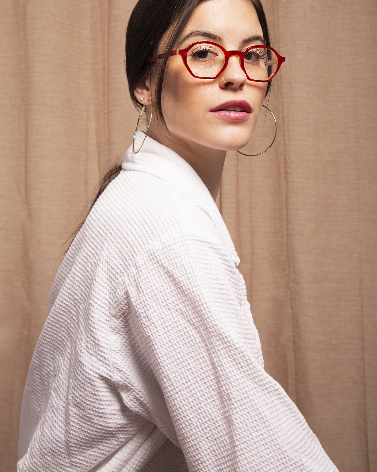Woman in a white waffle robe glancing over her shoulder, wearing red Kala eyewear glasses and gold hoop earrings