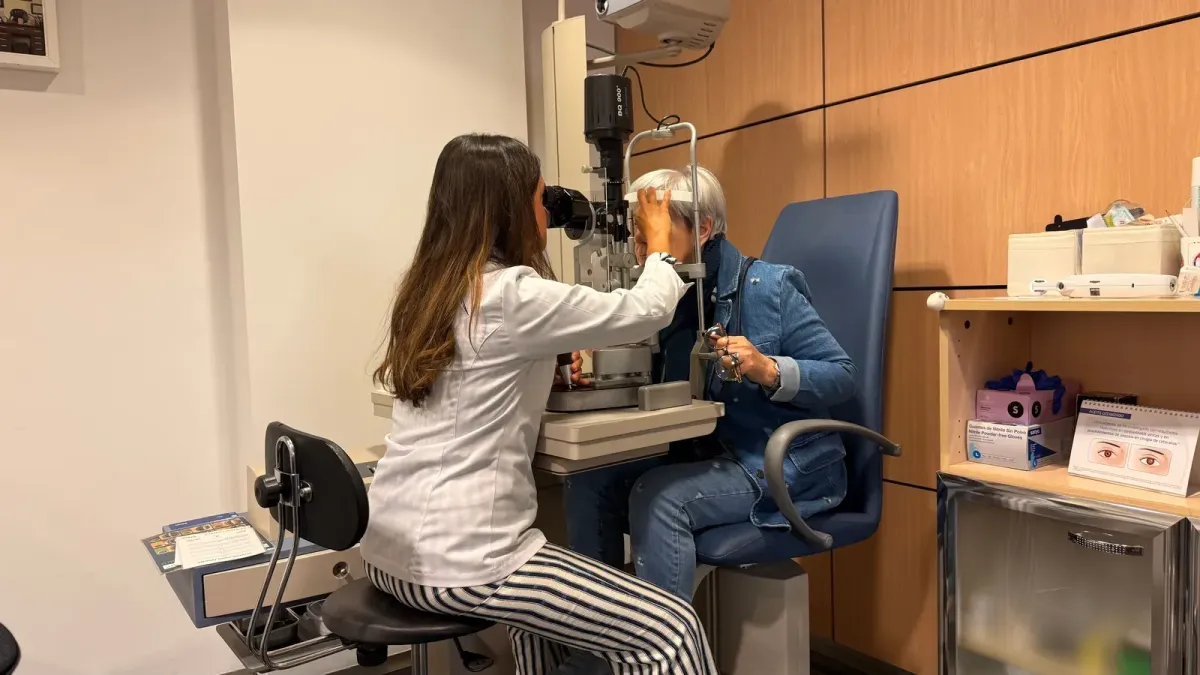 An optometrist uses a slit lamp to examine the eyes of an older adult patient during an eye exam in a clinic exam room.