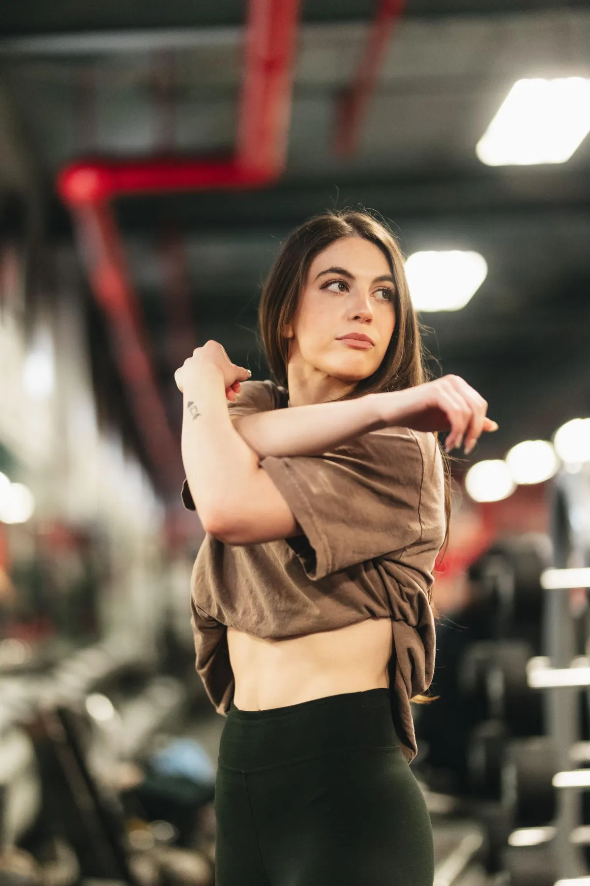 woman with long brown hair doing bicep curls in the gym, wearing black leggings and an oversized t-shirt, dumbbells lying on shelves behind her, photo taken from a distance, in the style of unsplash photography.