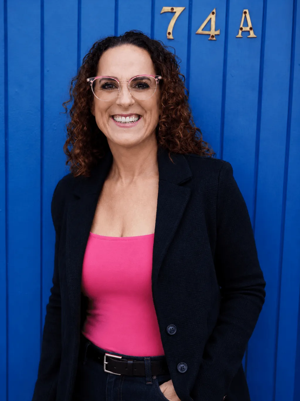 Louise, ethical leadership consultant, in a black blazer and pink top against a blue wall.