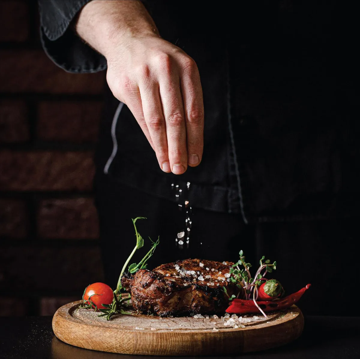 A chef's hand carefully sprinkles salt on a steak, a final touch that highlights the quality and craftsmanship of our kitchen.