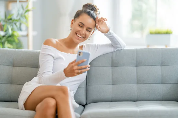 Lady in a white dress sitting on the couch, happily looking on her phone as she is making a life-changing income from the automated Done For You system.