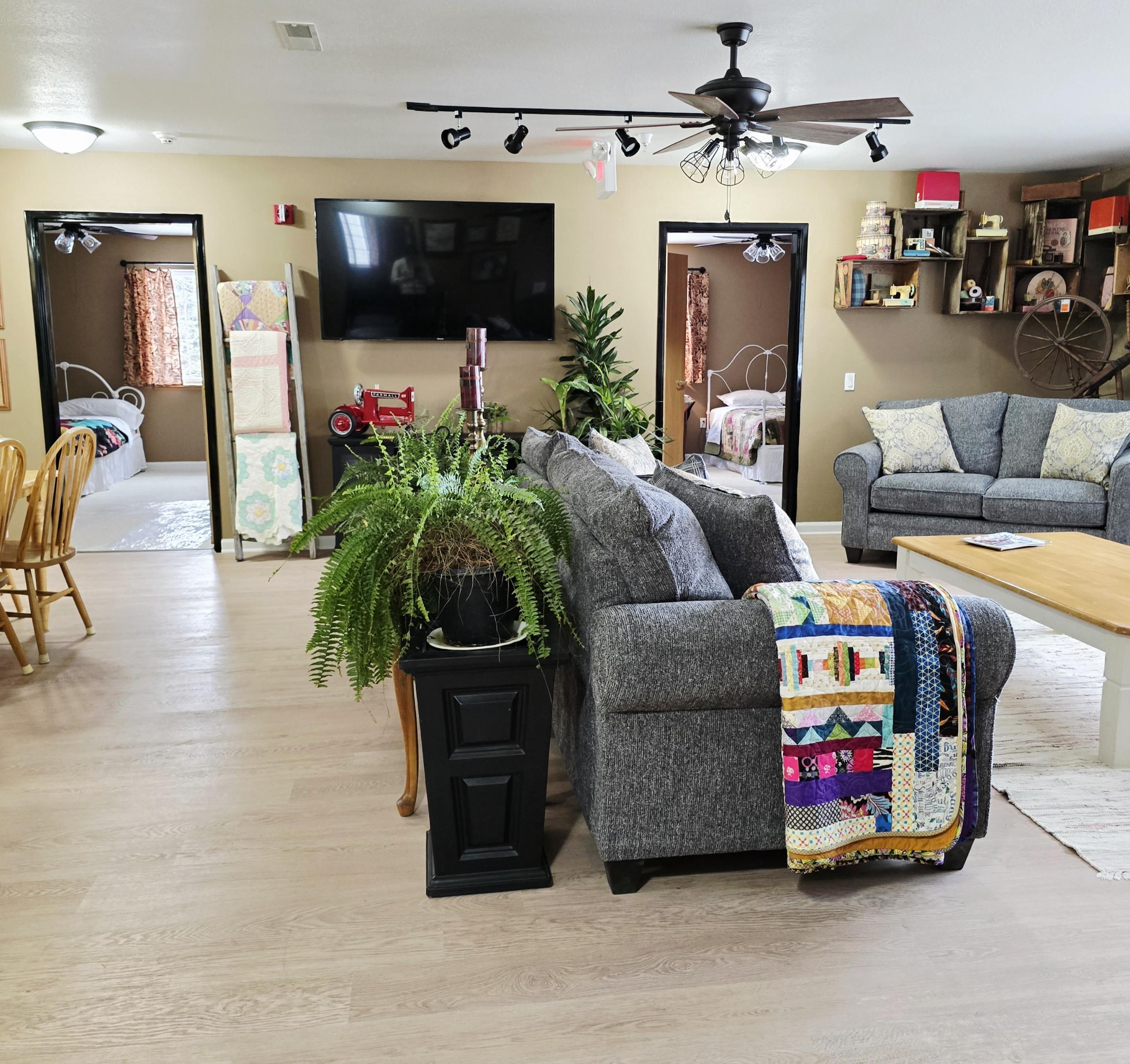 A group of women aged 40-75, gathered around a large wooden table, quilting together in a sunlit room with large windows overlooking a lush green landscape. The women are smiling, focused on their craft, and surrounded by colorful fabrics and sewing tools. The atmosphere is warm, inviting, and creative.