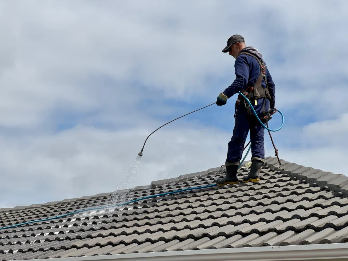 man on a brown/grey concrete tile roof applying a chemical cleaning solution from a battery powered hand held pump sprayer