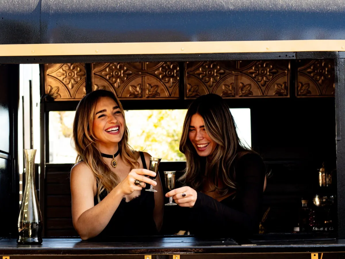 Two women laughing and toasting cocktails at the mobile bar trailer during a Denver cocktail catering event