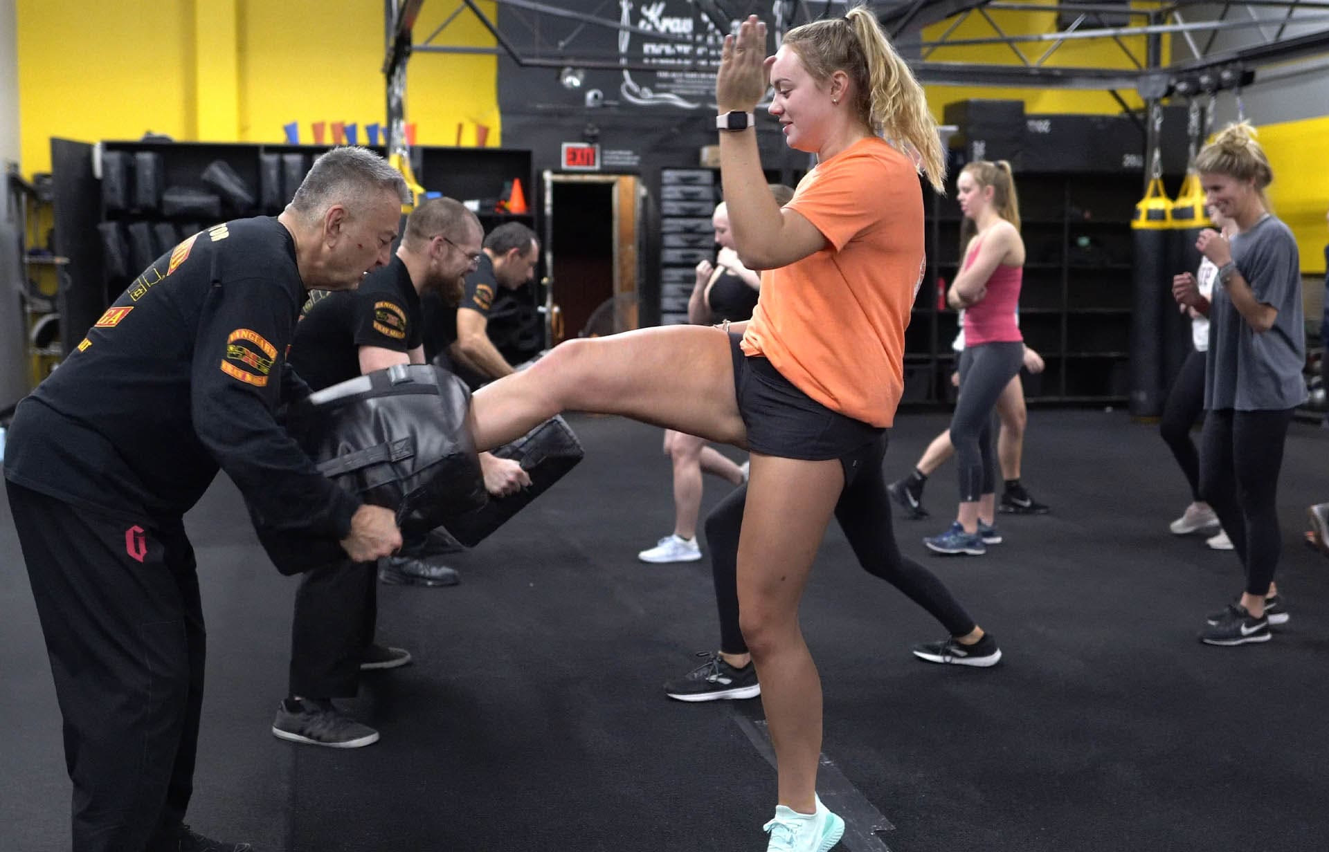 A woman in an orange shirt practices a high kick on a pad held by a martial arts instructor during a women's self-defense class inside a training gym, with other participants practicing in the background.