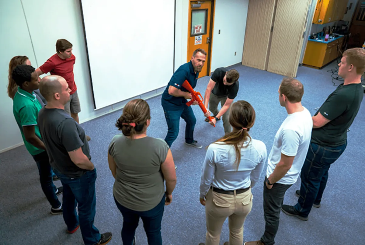 A safety instructor demonstrates a weapon disarm technique using a red training rifle prop while participants observe in a circle during an active shooter response training session.