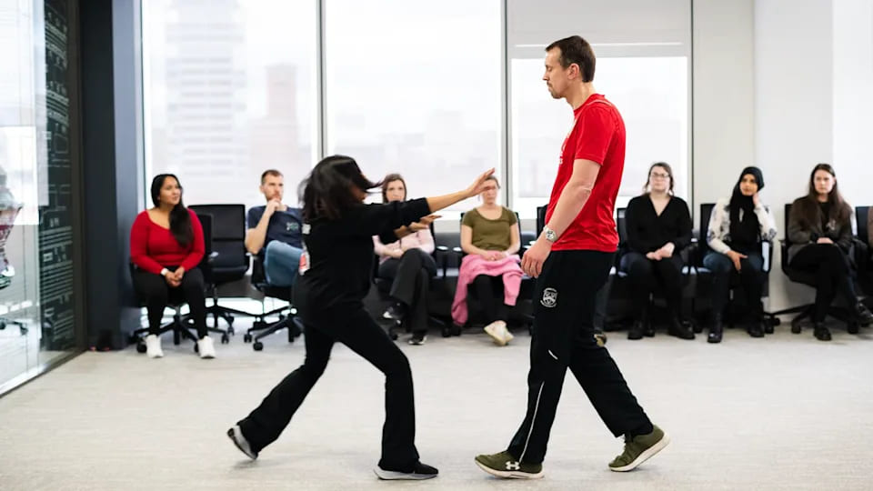 Two instructors demonstrating a self-defense scenario in a modern office space while employees watch