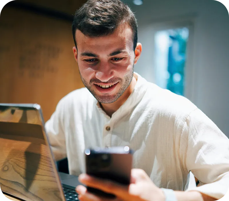 Man smiling while looking at his smartphone next to a laptop