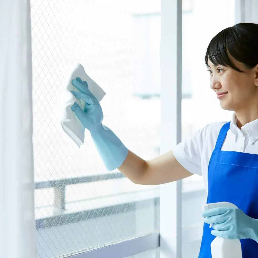 Professional maid in a blue apron and gloves wiping a glass window, representing standard home cleaning services by Maryland Spotless Maid Service.