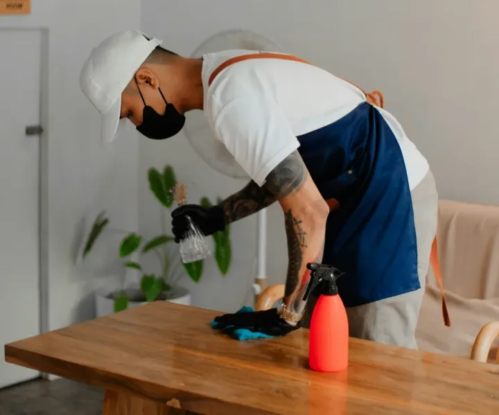 Professional background-checked cleaner wearing a mask and apron sanitizing a wooden table during a house cleaning service in Maryland.