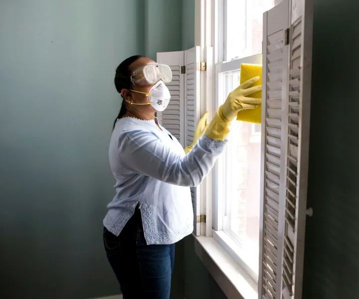 Professional background-checked cleaner wearing a protective mask, goggles, and yellow gloves cleaning a window and shutters during a residential service in Maryland