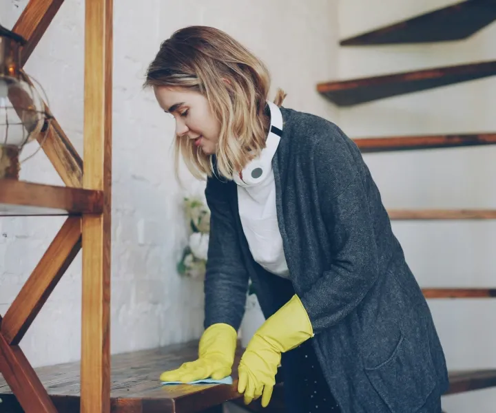 Professional background-checked cleaner wearing yellow gloves wiping and dusting a wooden shelf during a residential cleaning service in Maryland.