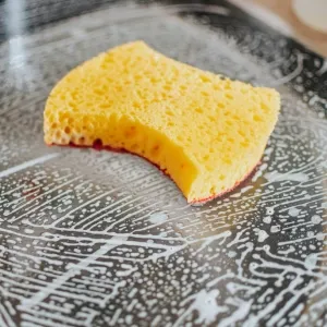 Yellow textured sponge scrubbing soap scum and heavy buildup on a glass surface, highlighting heavy duty cleaning services by Maryland Spotless Maid Service