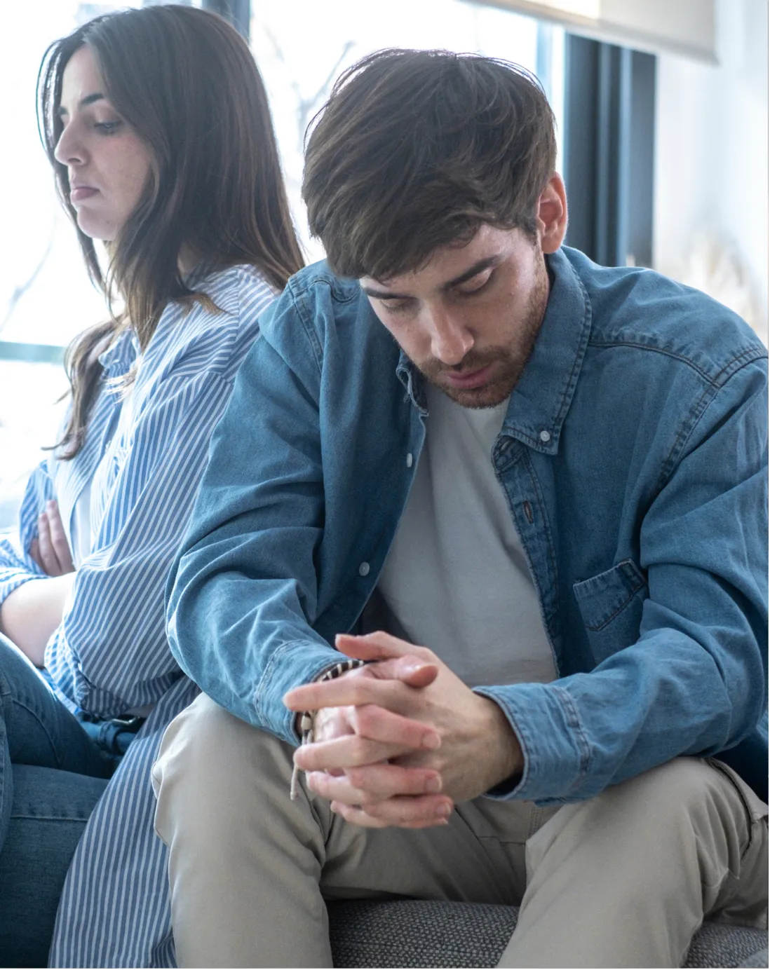 A man in a denim shirt sits with hands clasped, looking down, while a woman in a blue striped shirt sits behind him with her arms crossed indoors.