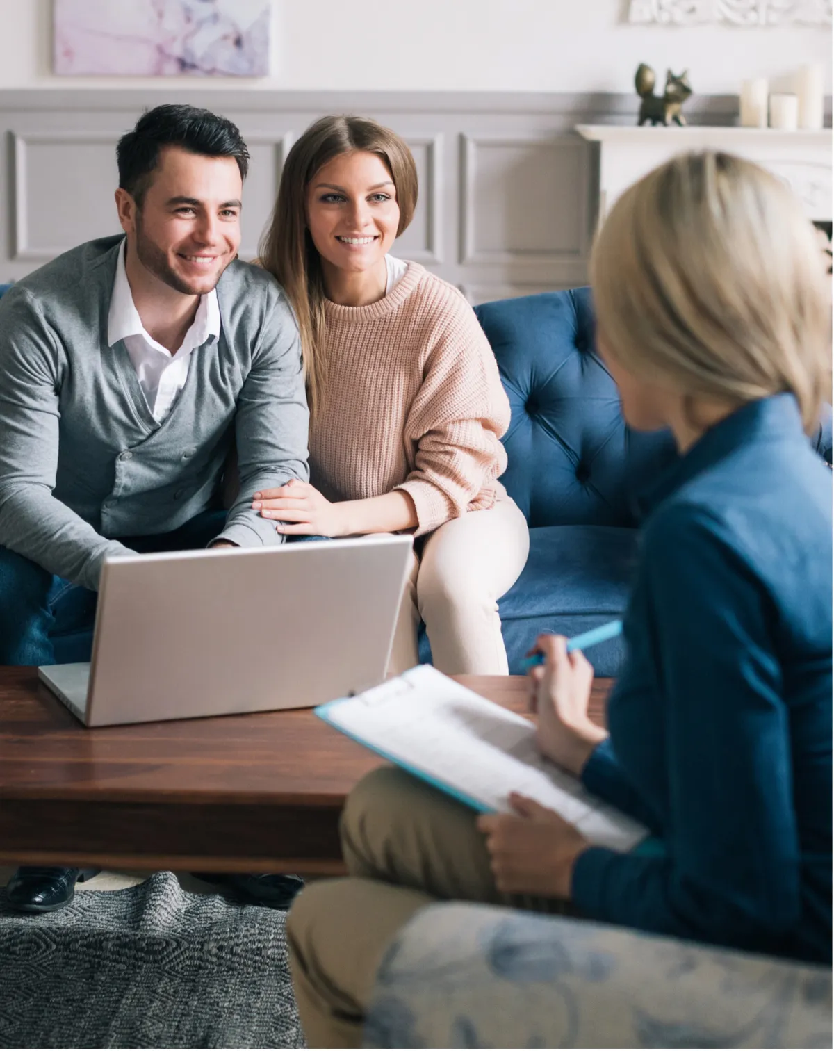 A smiling couple sits on a blue tufted sofa with a laptop, consulting with a professional who holds a clipboard in a bright, modern living room.