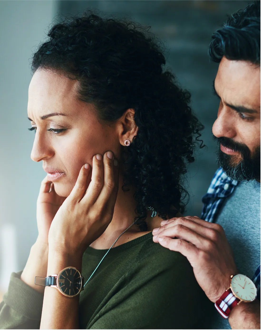 A man with a beard gently touches the face and shoulder of a woman with curly dark hair, both wearing wristwatches in a close-up indoor portrait.
