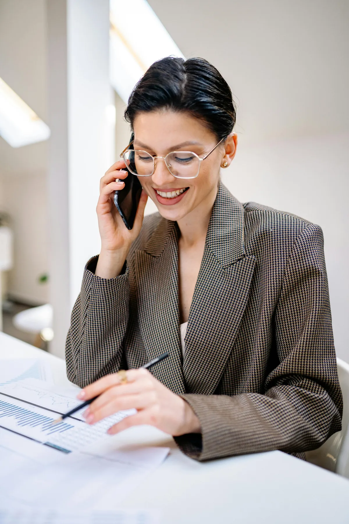 A woman with short dark hair and glasses holding a phone to her ear and writing notes with a pen. 