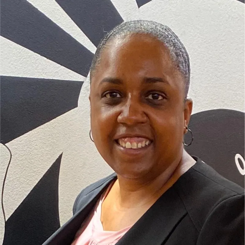 Smiling woman with short gray hair wearing a black blazer over a pink top, standing in front of a black-and-white mural.