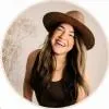 Smiling woman with long dark hair wearing a wide-brimmed brown hat in a warm, softly lit portrait.