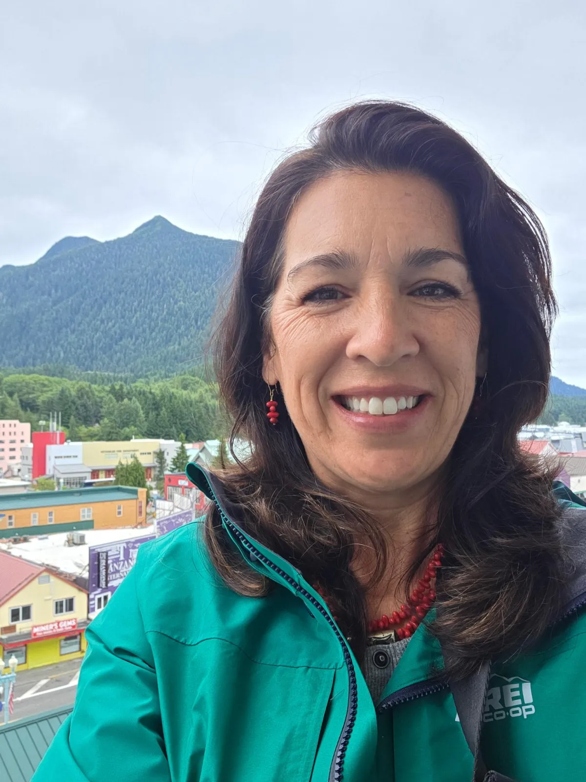 Smiling woman with long dark hair wearing a teal jacket, photographed outdoors with a mountain and small town in the background.