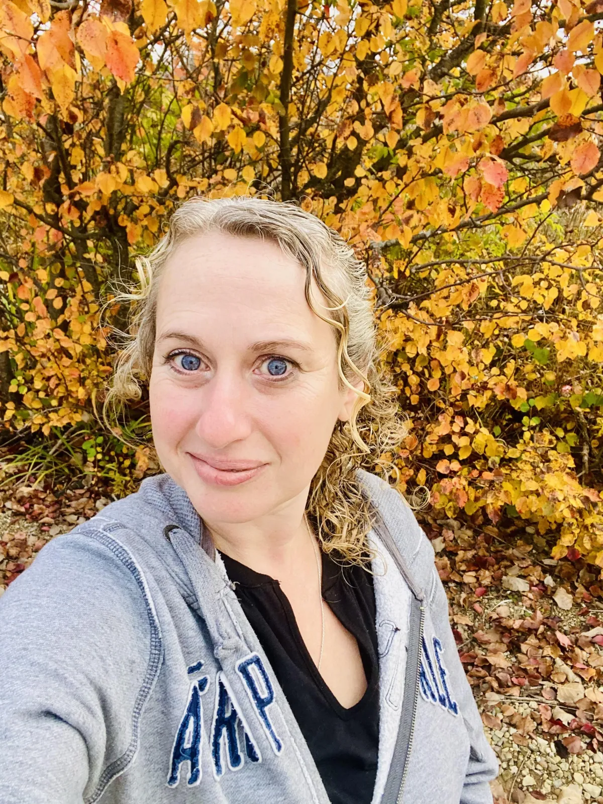 Smiling woman with curly blonde hair wearing a gray hoodie, photographed outdoors in front of vibrant golden and orange autumn foliage.