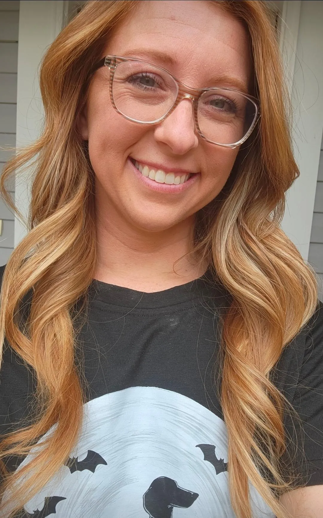 **Alt text:** Smiling woman with long red-blonde hair and clear glasses wearing a black T-shirt, photographed outdoors in natural light.