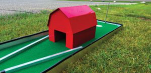 Children at a backyard birthday party, each taking turns at a colorful mini golf hole. Balloons and party decorations surround the area, and parents cheer from the sidelines. The scene is lively and festive.