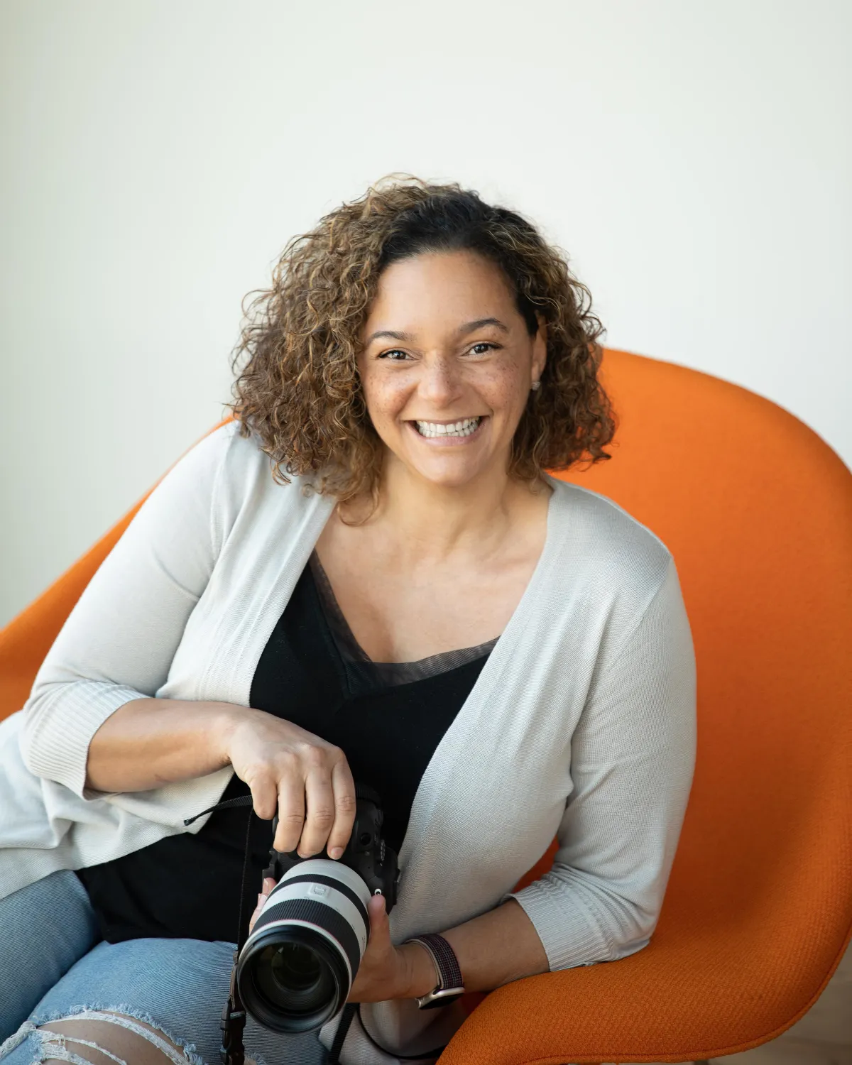 Smiling woman with curly hair wearing a light cardigan, sitting on an orange chair in a bright studio. Professional headshot by Los Angeles portrait photographer Kendra Greenberg Photography.