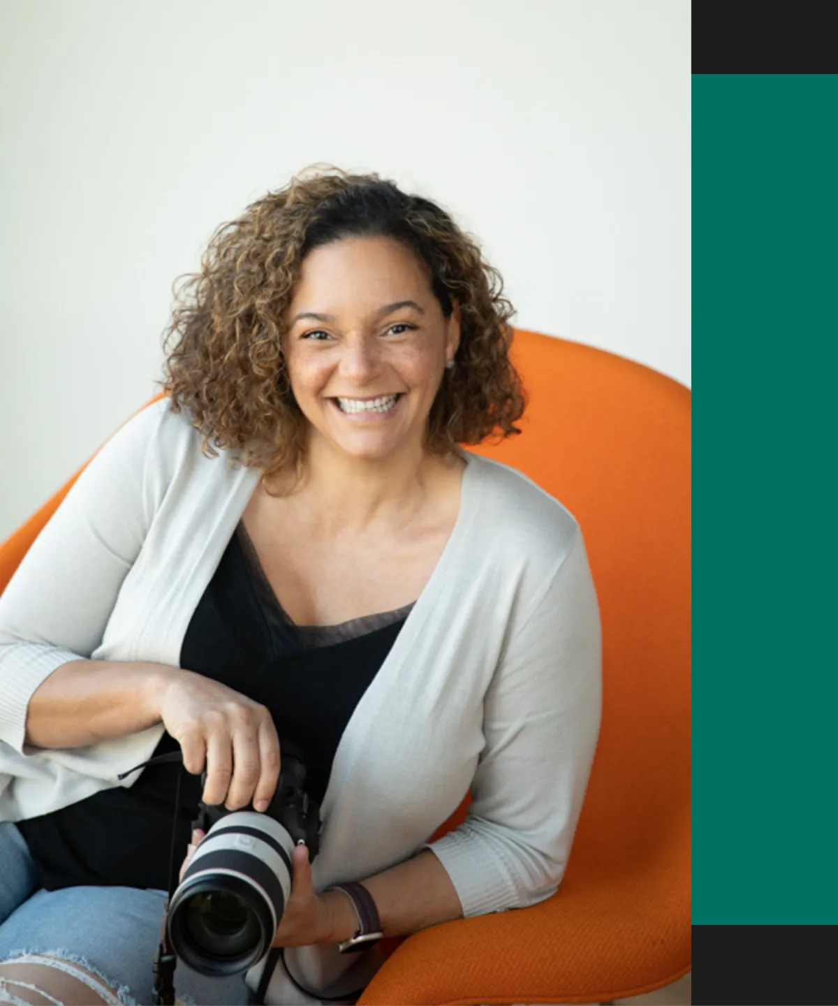 Smiling woman with curly hair wearing a light cardigan and black top, sitting against an orange chair. Photograph by Los Angeles photographer Kendra Greenberg Photography.