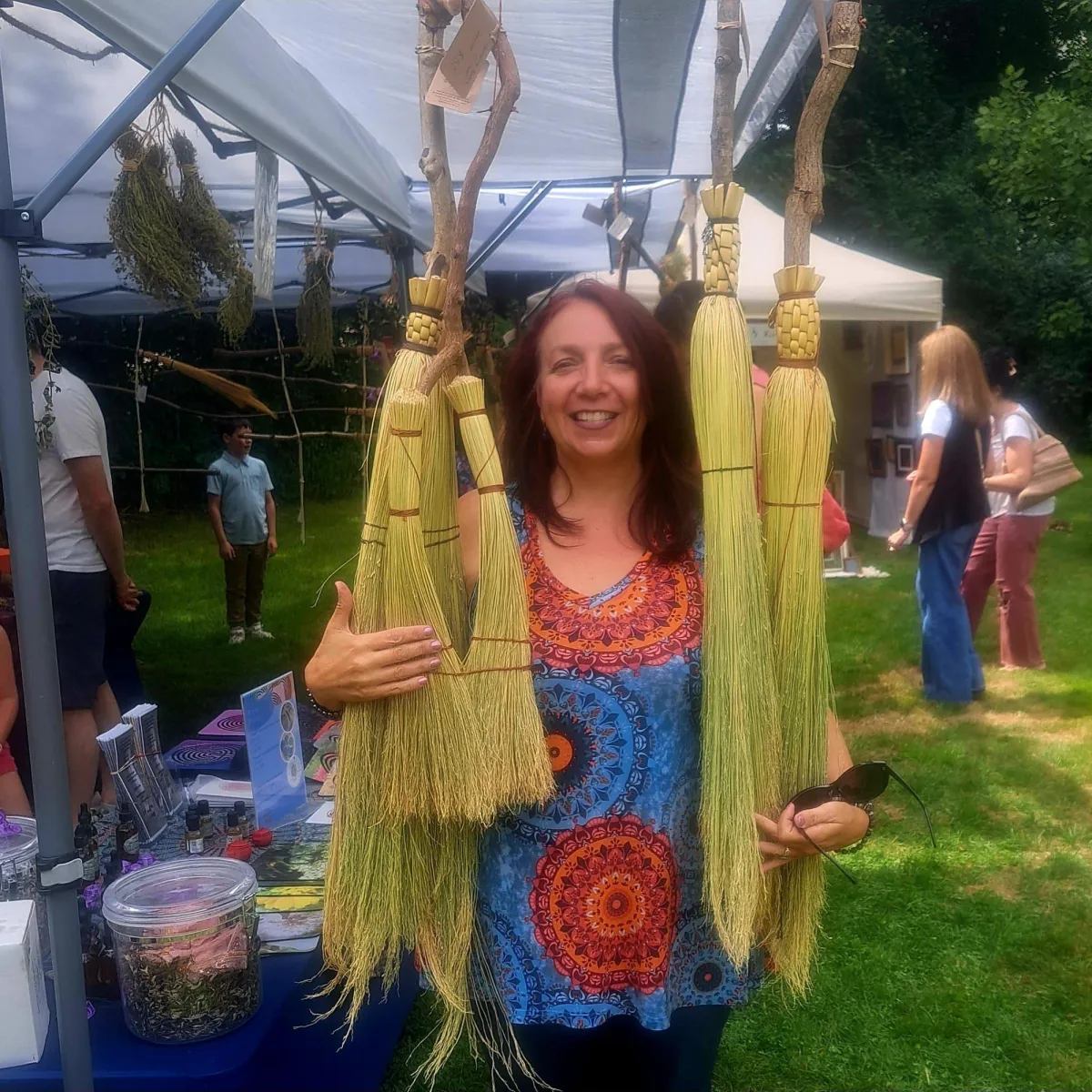 A portrait of a middle-aged artisan with gentle features, wearing a linen apron and holding a finished broom, standing in a sunlit studio filled with natural materials and tools. The instructor exudes warmth, expertise, and a welcoming presence.