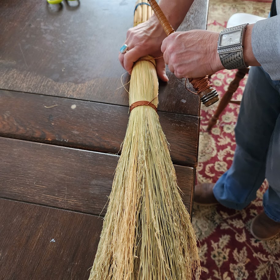 A close-up of a skilled artisan's hands weaving natural straw into a traditional broom, with wooden handles and colorful twine, set against a softly blurred workshop background. The image captures the intricate craftsmanship and tactile process, evoking a sense of heritage and creativity.