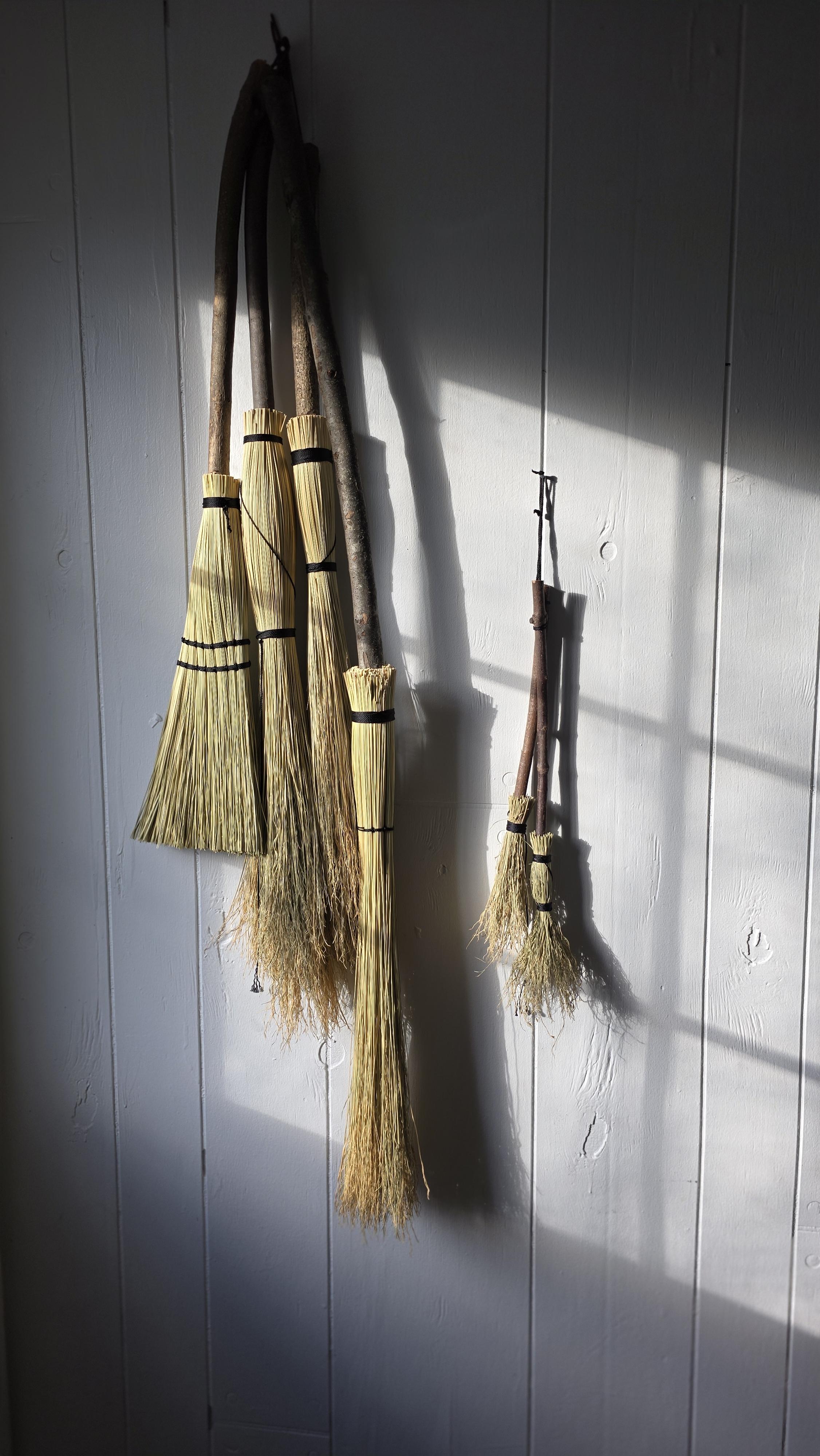 A vibrant workshop scene showing a diverse group of adults gathered around a rustic table, each working on their own broom project, with tools, straw, and wood scattered about. The atmosphere is collaborative, creative, and filled with focused energy.