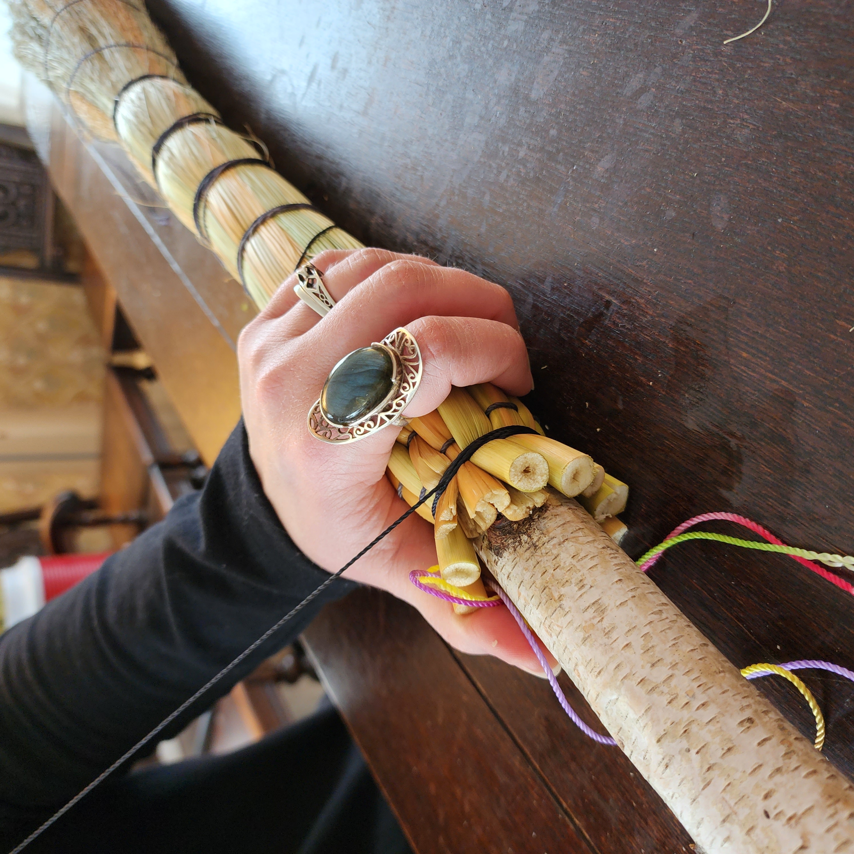 A close-up of a skilled artisan's hands weaving natural straw into a traditional broom, with wooden handles and colorful twine, set against a softly blurred workshop background. The image captures the intricate craftsmanship and tactile process, evoking a sense of heritage and creativity.