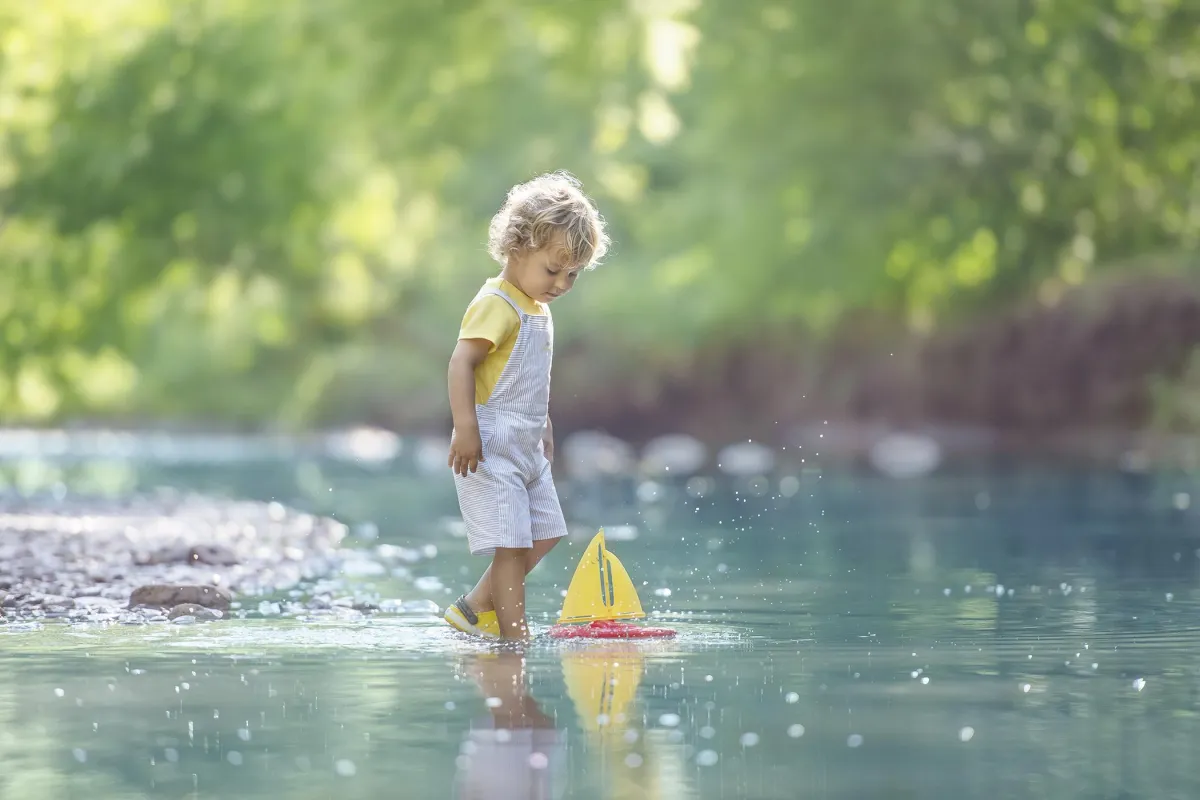 Photo of the little boy playing with his ship toy by the shore of the river in the morning light. 