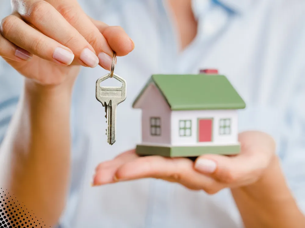 Hand holding a key to a small model house, symbolizing a new home purchase.
