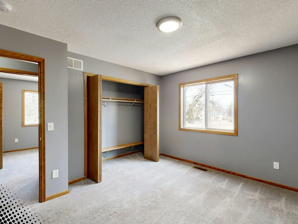 Empty bedroom with beige carpet and natural light coming from a window, featuring a simple design and an unoccupied closet