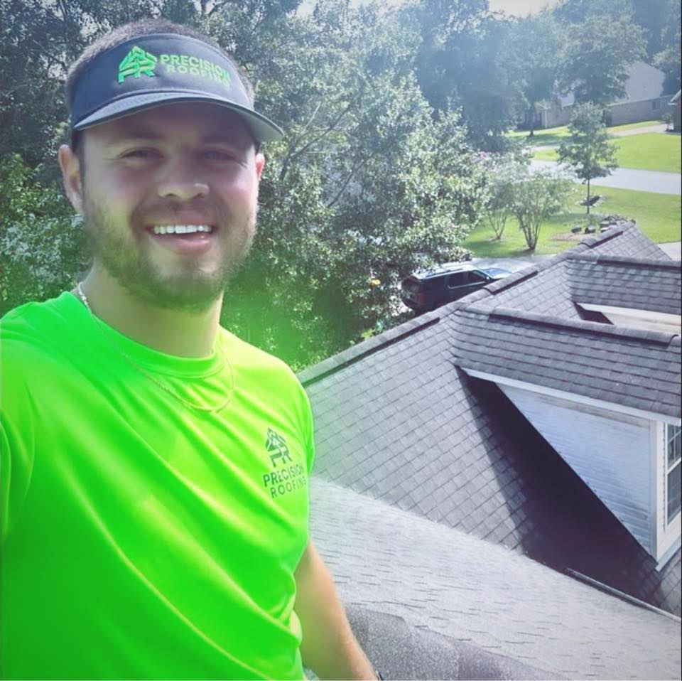 Smiling roofing professional in a bright green shirt with "Precision Roofing" logo, standing on a sloped roof, showcasing roofing work and surrounding residential area.
